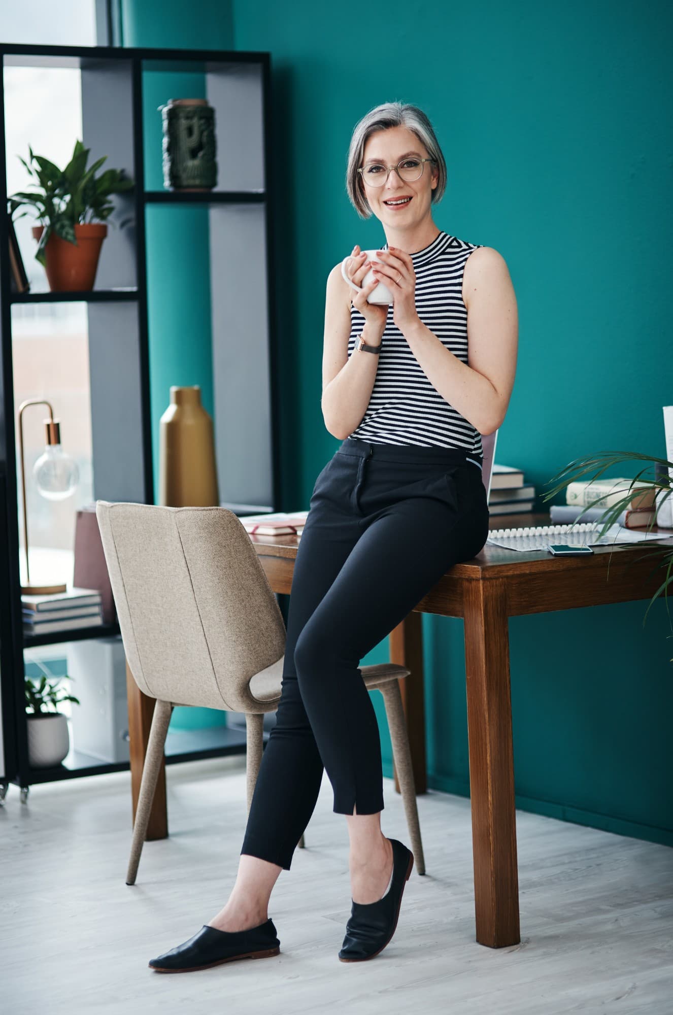 A woman with short grey hair and glasses, smiling, holding a mug of coffee and leaning on a desk in a teal office.